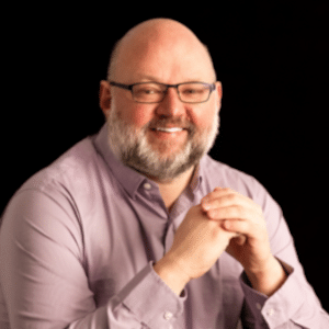 A portrait of Aaron DeVries. He is a bald man with a two-toned grey beard and glasses. He is wearing a lavender collared shirt. The background is dark and he is strongly lit from his left side.