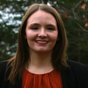 A portrait of Haley Clark. She has parted dark hair and is wearing an orange shirt with a pleated neck and a black cardigan. Behind her are some autumn trees.