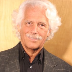 A portrait of Steven Schwartz. He has a white mustache and ear-length curly white hair brushed back on his right, falling over his forehead on his left. He is wearing a brown blazer with a slight plaid and a brown open-collared shirt.